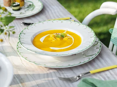 Une assiette à soupe Colourful Spring avec une soupe de patates douces et de mangue, garnie d'herbes et de crème, est posée sur une table décorative en bois, à côté de laquelle se trouve une fourchette.