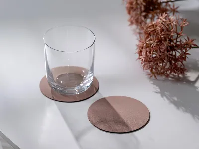 Un bicchiere trasparente si trova su un sottobicchiere rotondo accanto a un altro sottobicchiere su una superficie bianca con fiori secchi nelle vicinanze.