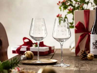 Two Villeroy & Boch Toy’s Delight glasses on a wooden table with Christmas presents, a bottle of wine, a pine cone, and festive decorations in the background.