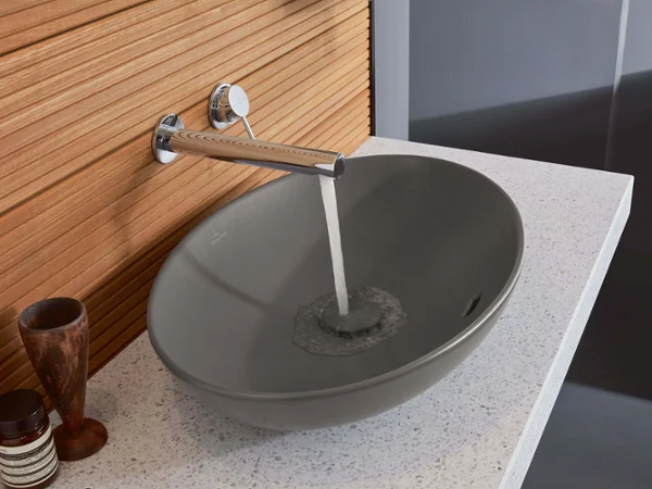 Grey Loop & Friends washbasin in front of a wooden wall Modern bathroom with a grey Loop & Friends surface-mounted washbasin and a wood-accented wall tap with running water.
