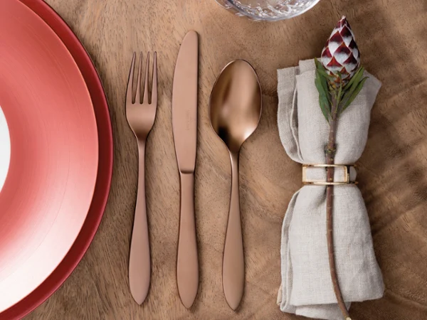 manufacture-copper-flatware-on-wood-table A place setting with a red plate, rose gold fork, knife, and spoon, a glass, and a napkin with a floral napkin ring on a wooden table.