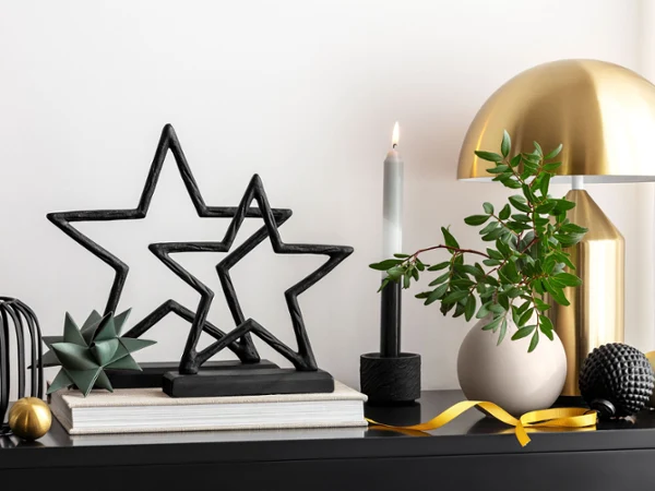 Decoración de mesa con figuras de estrellas Black XMAS, una lámpara dorada, libros, una vela encendida y diversos objetos decorativos.