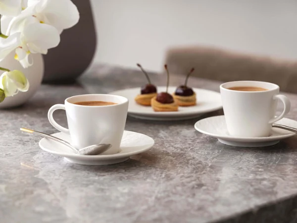 Two Manufacture espresso cups and a plate of pastries on a marble table with orchids in the background.
