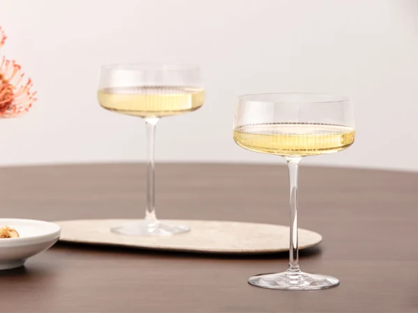 Zwei gefüllte Champagnergläser der Afina Kollektion auf einem Holztisch, daneben eine kleine Schale mit Snacks und eine Blumenvase mit orangefarbenen Blüten.