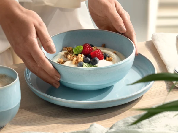 Hands placing a blue mother-of-pearl aqua bowl with yoghurt, muesli, blueberries and raspberries on a blue plate.