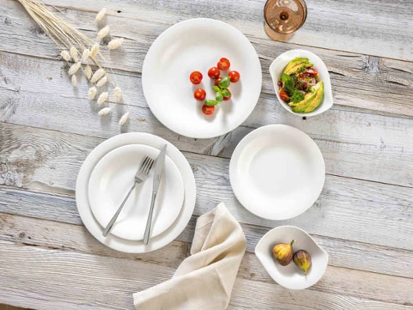 Vajilla Rose Cottage sobre una mesa rústica de madera Una mesa de estilo rústico con platos Essential White y New Cottage, tenedor y cuchillo, una servilleta beige y cuencos con tomates cherry, rodajas de aguacate e higos. Arriba a la derecha hay un vaso de vino rosado.