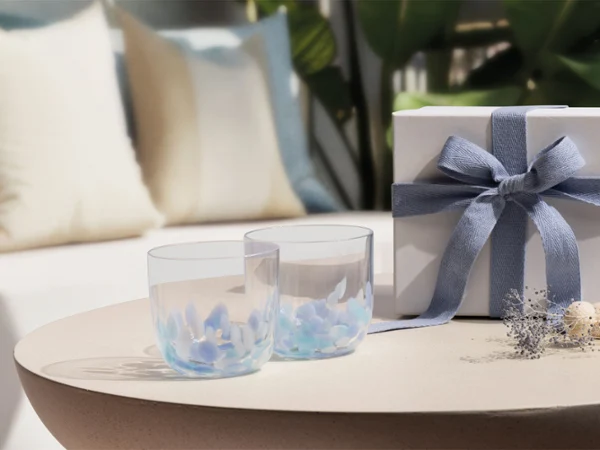 Dos vasos transparentes Like Aqua y una caja de regalo blanca. Dos vasos transparentes Like Aqua y una caja de regalo blanca con un lazo azul sobre una mesa redonda; sofá y cojines al fondo.