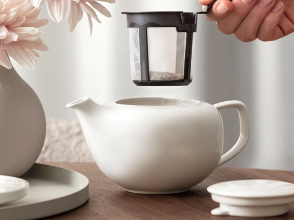 Tea Preparation with Afina A hand gently holds a tea filter over a white Afina teapot. The teapot stands on a wooden table, with a flower vase completing the scene in the background.