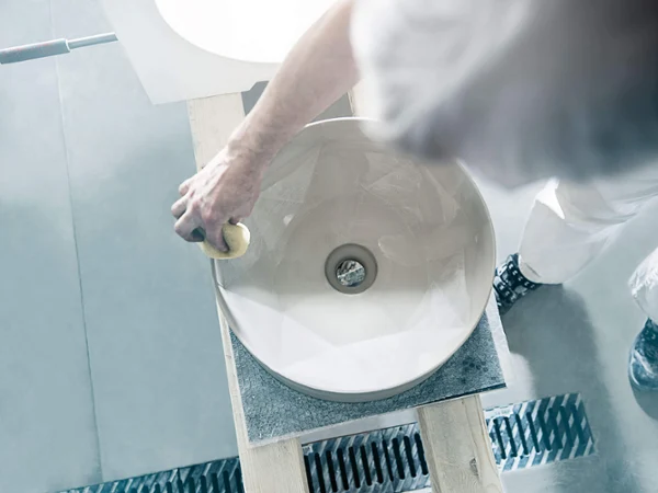 Czyszczenie umywalki Villeroy & Boch Osoba czyszcząca gąbką w warsztacie białą ceramiczną umywalkę. Umywalka stoi na drewnianej konstrukcji, a przedstawiona osoba ma na sobie białe ubranie i czarne buty.