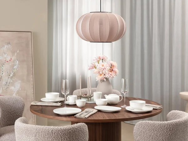 Stylish enjoyment with Afina A modern dining room with a round wooden table, white Afina tableware, Afina glasses, pink flowers and a beige pendant lamp.