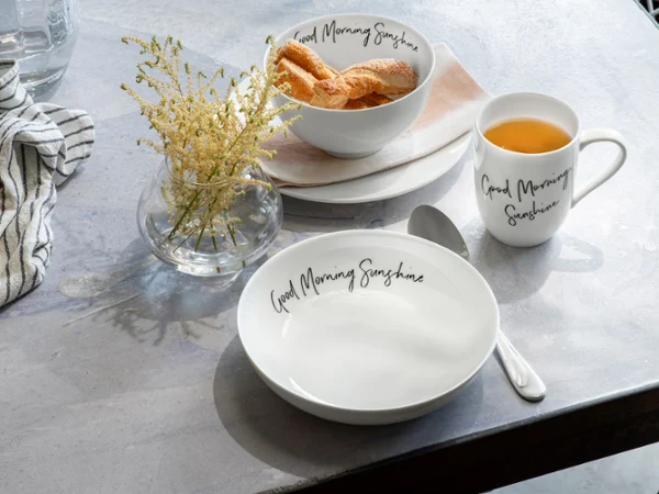 Taza, plato y bol Statement "Good Morning Sunshine" sobre una mesa Una mesa con una taza de café y un plato con croissants de Villeroy & Boch.