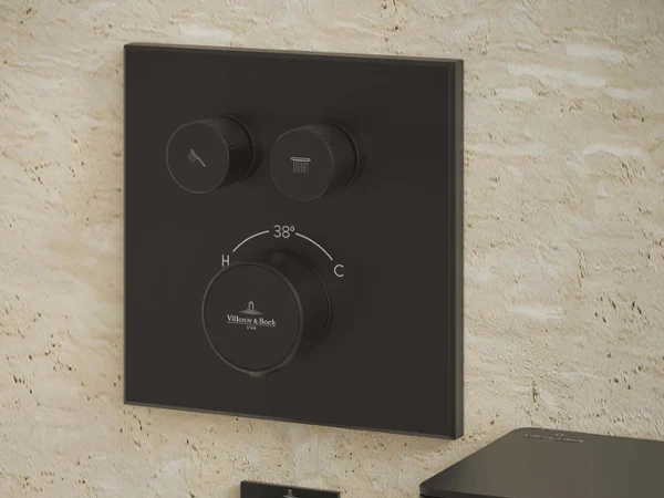 Thermostat encastré noir et flexible de douchette montés sur un mur de salle de bain carrelé beige.