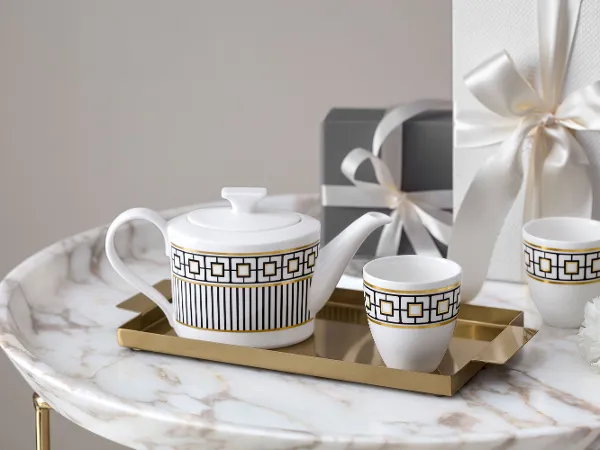 A white and gold patterned teapot and cups from MetroChic on a golden tray on a marble table. Two wrapped presents and a small white flower can be seen in the background.