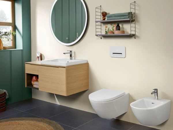 Łazienka z wyposażeniem Subway 3.0 obejmującym białą miskę WC z bidetem, umywalkę i szafkę podumywalkową w wersji Oak Nowoczesna łazienka z umywalką ścienną i drewnianą szafką podumywalkową, okrągłym lustrem, toaletą wiszącą, bidetem i metalową półką ze złożonymi ręcznikami i dekoracjami. Ciemne płytki podłogowe i zielone ściany.