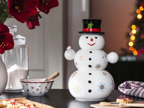 Un bonhomme de neige Christmas Toys Memory avec un plat creux Toy’s Delight Un bonhomme de neige Villeroy & Boch avec un chapeau noir et un ruban rouge se tient sur une table à côté d’un sucrier, d’un bol mélangeur et de fleurs rouges. En arrière-plan, des lumières de Noël sont floues.