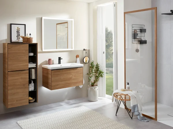 Drewniane meble łazienkowe z kolekcji Subway 3.0 Nowoczesna łazienka z drewnianą szafką z umywalką, szafkami ściennymi, prysznicem zlicowanym z podłogą ze szklaną ścianką i dużym oknem zapewniającym naturalne światło. Na podłodze leży dywan, a na małym taborecie znajdują się przybory do kąpieli.