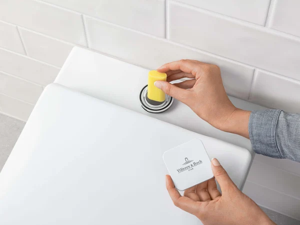 Una persona arroja una piedra sanitaria de color amarillo a la cisterna de un retrete. Al fondo se ven paredes de azulejos blancos.