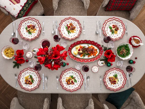 A festive dining table setting A festive dining table setting for eight people with red and white patterned plates, red flowers, glasses, cutlery and various dishes, including pasta and salad, on a white tablecloth.