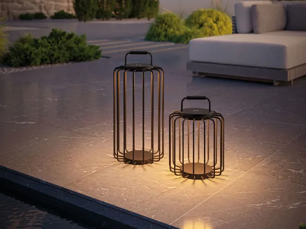 Zwei moderne Laternen mit warmem Licht, die abends auf einer Terrasse neben einem Gartensofa platziert werden.