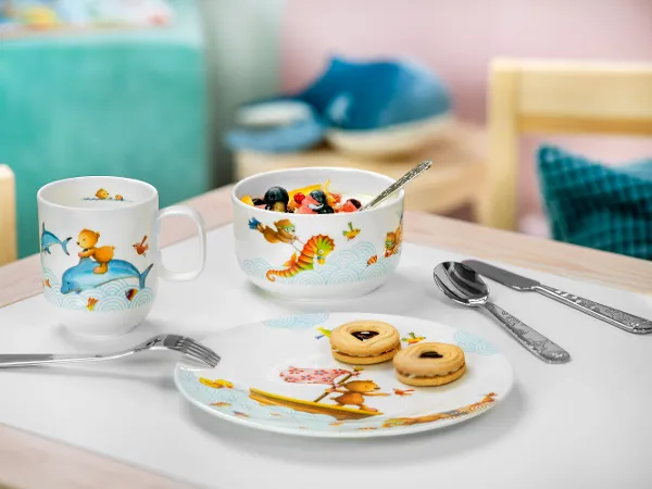 De la vaisselle pour enfants Happy as a bear avec de la salade de fruits et des biscuits Une présentation de petit-déjeuner avec une salade de fruits, des biscuits sur une assiette et deux tasses de vaisselle pour enfants Happy as a bear sur une table.