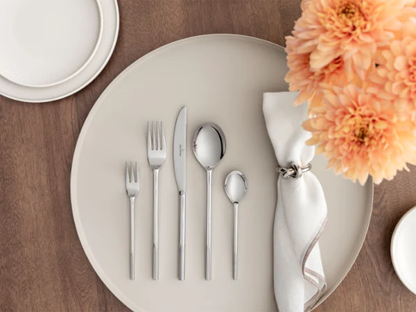 Gedeckter Tisch mit fein säuberlich auf einem großen weißen Afina Teller angeordnetem Silberbesteck, einer gefalteten weißen Serviette mit Ring, einer Teilansicht von orangefarbenen Blumen und zwei kleinen Tellern auf einem braunen Tisch.