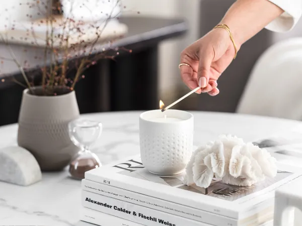 A person lighting a Manufacture scented candle in white on a coffee table with decorative objects and books.