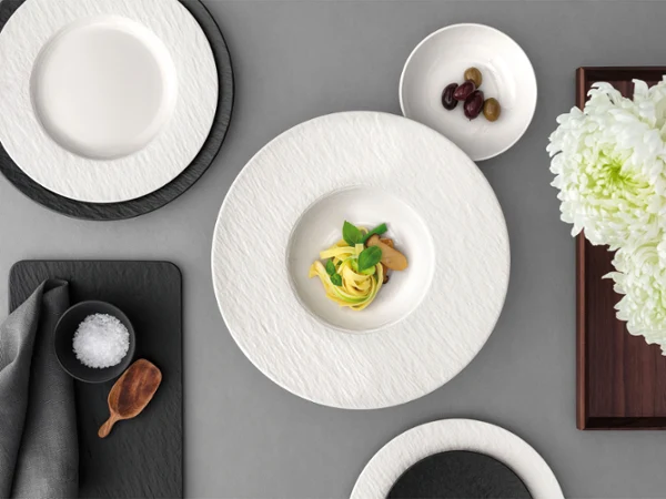 A formal table setting with a Manufacture pasta plate A formal table setting with a white past plate with food in it on a grey table.