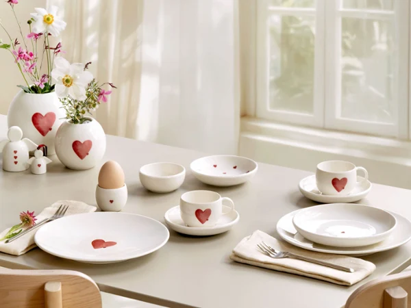 Vajilla y artículos decorativos de with love en la mesa La mesa del desayuno está puesta con vajilla blanca con motivos de corazones rojos de with love, flores en jarrones de with love y utensilios cuidadosamente dispuestos.