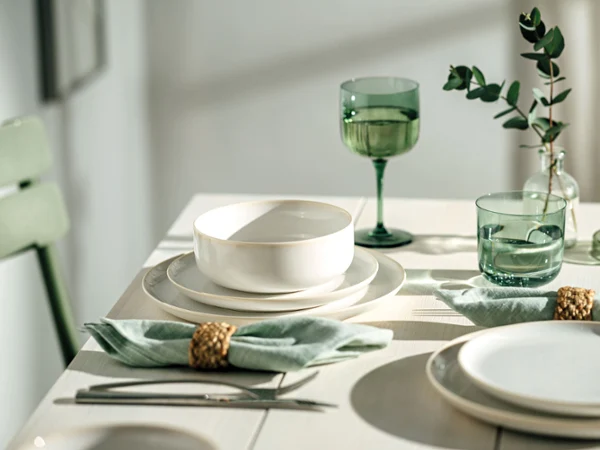 Platos artesanales blancos con vasos Like verdes sobre una moderna mesa de madera Una mesa de comedor con platos blancos Crafted, vasos verdes Like Glass y cuencos, además de servilletas verdes con soportes dorados y adornos de hojas verdes.