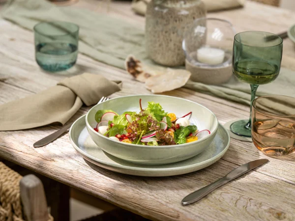 A Perlemor bowl of salad sits on a set dining table A bowl of salad sits on a set dining table with Perlemor glasses, plates, and beige napkins.