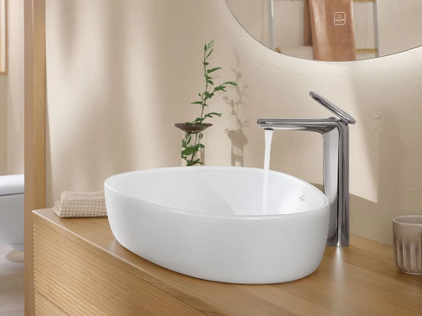Mobile lavabo con lavabo ovale bianco della collezione Antao ed elegante rubinetto cromato con acqua corrente. Un asciugamano beige piegato e una pianta verde si trovano sulla mensola di legno accanto al lavabo.