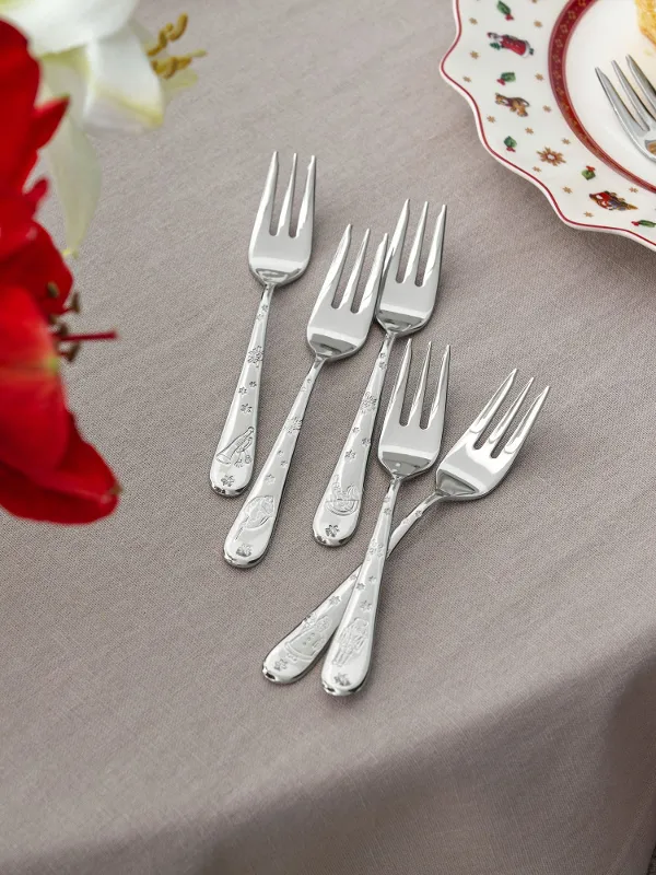 Five dessert forks on a beige tablecloth next to a plate with cake and whipped cream and a vase with red flowers.
