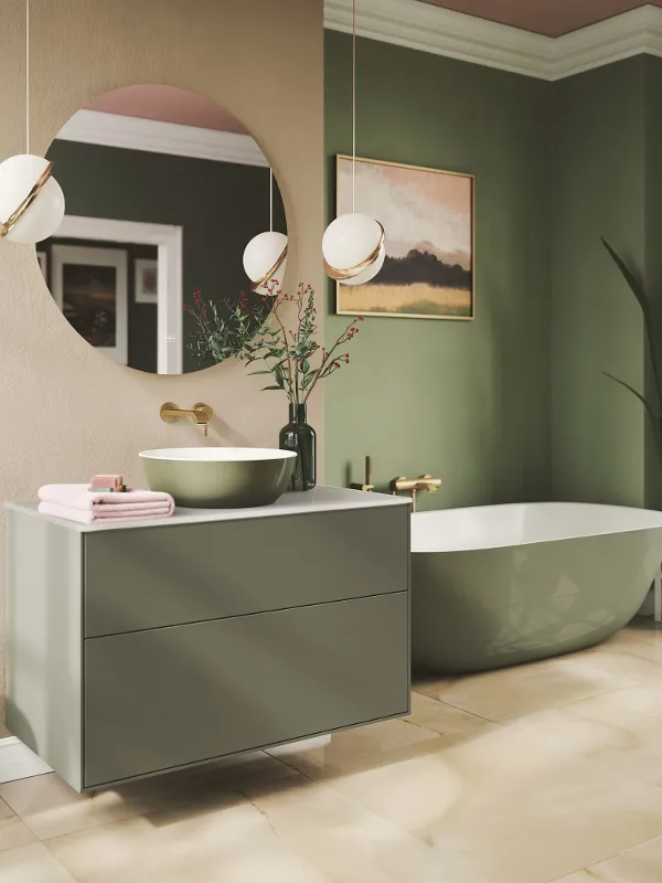 Bagno verde creativo con lavabo Artis di Villeroy & Boch Bagno moderno con lavabo e vasca verdi, specchio rotondo, rubinetto a parete, lavabo bianco, piante ornamentali e due lampade a sospensione. Sullo sfondo si vedono una vasca da bagno e una parete artistica.