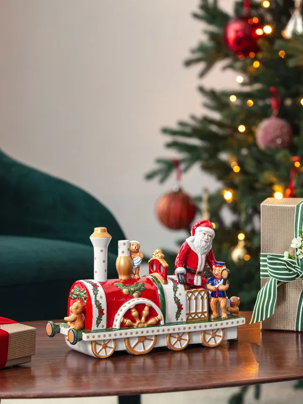  decorated Christmas tree, presents, and a Christmas Toys Memory locomotive on a wooden table next to a green chair.