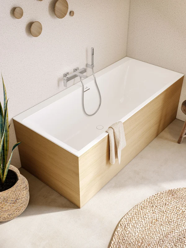 Nowoczesna łazienka z wanną prostokątną z kolekcji Subway marki Villeroy & Boch Nowoczesna łazienka z wanną prostokątną z kolekcji Subway marki Villeroy & Boch, drewnianym stołkiem z rośliną, tkanym dywanikiem i dużą rośliną doniczkową obok wanny. Ściany mają jasne cętkowane wykończenie z okrągłymi drewnianymi akcentami.