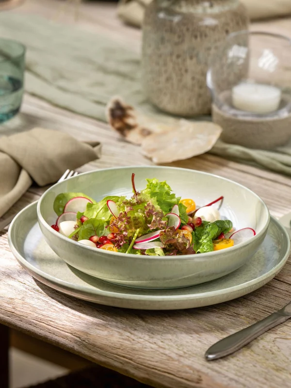 Eine Salatschüssel steht auf einem gedeckten Esstisch mit Perlemor-Gläsern, Tellern und beigen Servietten.