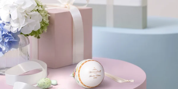 A pastel pink gift box with a white bow sits on a pink table next to the white and gold Annual Easter Ornament, with a bow, light green sweets and a vase with white and blue flowers. A blue gift box stands in the background.