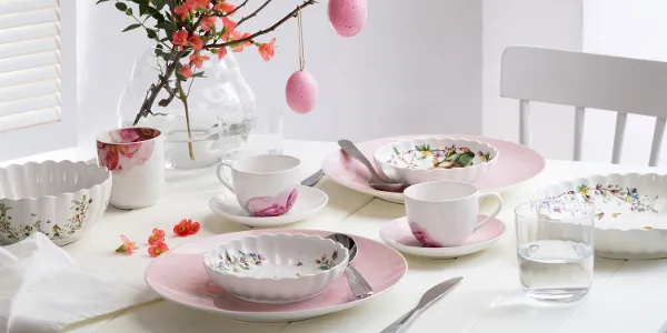 A bright, springtime dining table set with pastel-colored dinnerware from the Rose Garden and Spring Fantasy collections, decorated with floral patterns and Easter eggs hanging from a branch as a centerpiece.