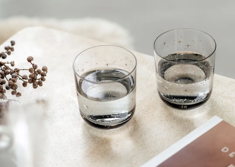 Two glasses of water on a textured beige surface with dried flowers and a book next to them.