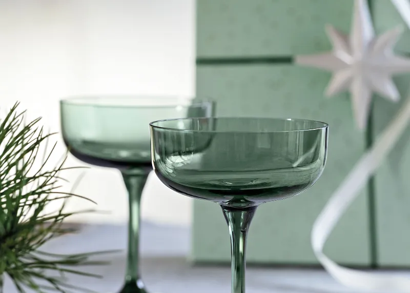 Like Sage glass ideal as a gift Two green cocktail glasses on a white surface with a green gift box and fir branches in the background.