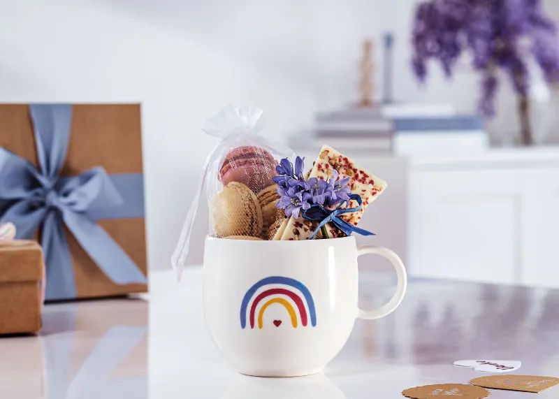 Une tasse « With Love » de Villeroy & Boch au design arc-en-ciel remplie de macarons Une tasse « With Love » de Villeroy & Boch au design arc-en-ciel, remplie de macarons, d’une tablette de chocolat et avec fleurs violettes sur une surface blanche avec des boîtes cadeaux et des livres en arrière-plan.