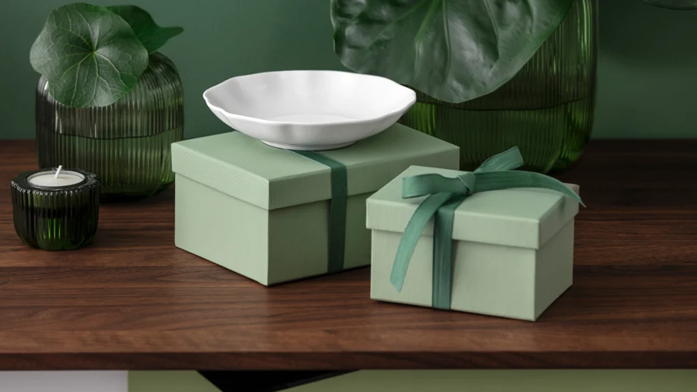 Two green gift boxes stand on a wooden surface with a white Pura bowl. Two green gift boxes, one of them with a bow, stand on a wooden surface with a white Pura bowl and green glass decorations.