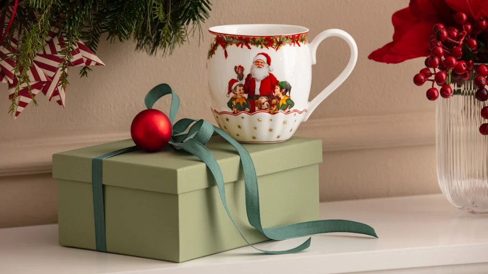 Annual Chistmas edition 2026 mug A green gift box with a bow, a red Christmas ornament, and a mug with a Santa Claus motif from Villeroy & Boch in front of a white background, with Christmas decorations visible in the background.