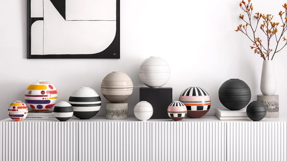 A series of patterned spherical tableware sets on a white shelf, underneath a geometric black-and-white painting featuring a vase with orange flowers.