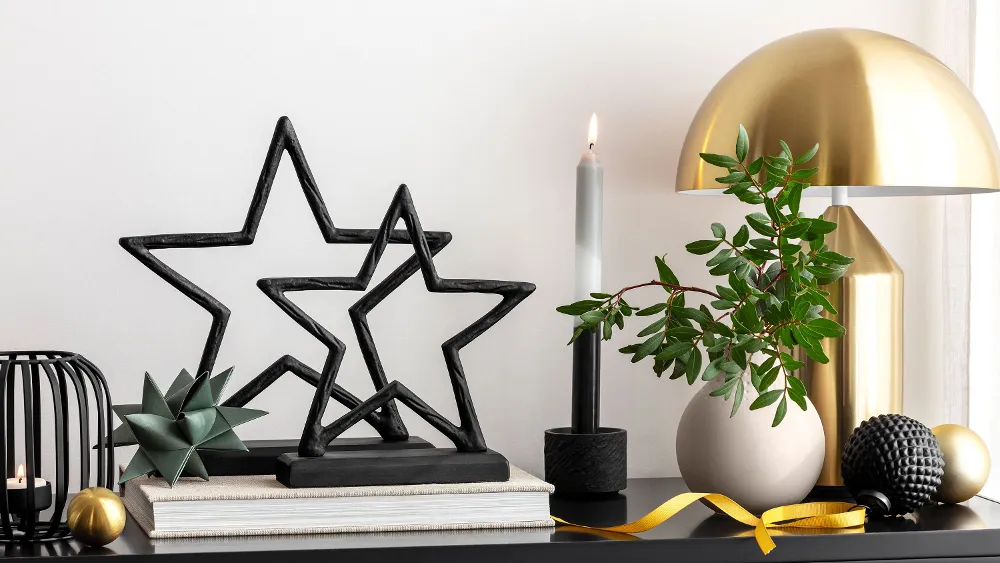 New Year decorations with products from the Villeroy & Boch Black Xmas series Table decoration with black Black Xmas star sculptures, a gold lamp, books, a lit candle and various decorative objects.
