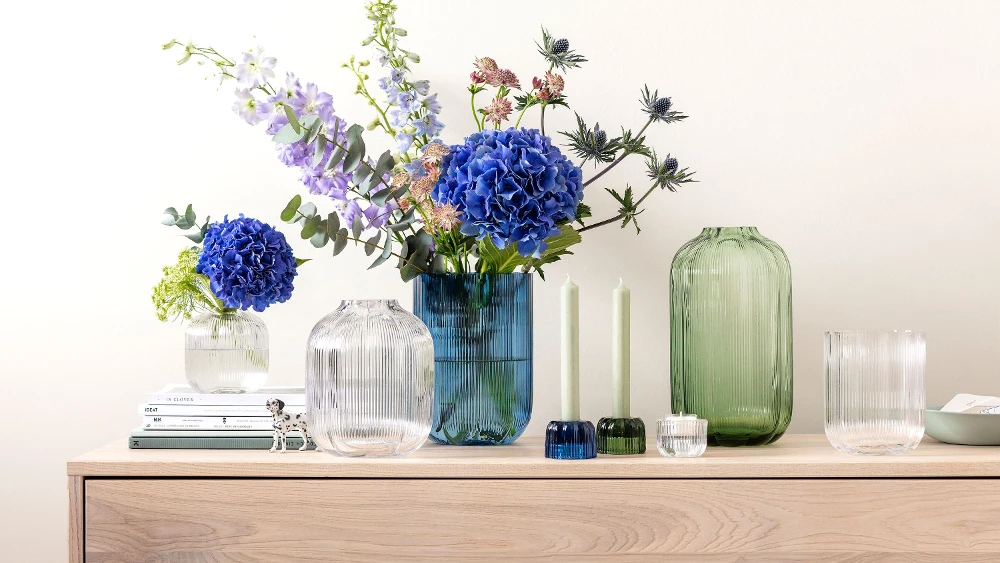 A sideboard with Fleur vases from Villeroy & Boch, decorated with blue and purple flowers, next to two green candles and stacked books.