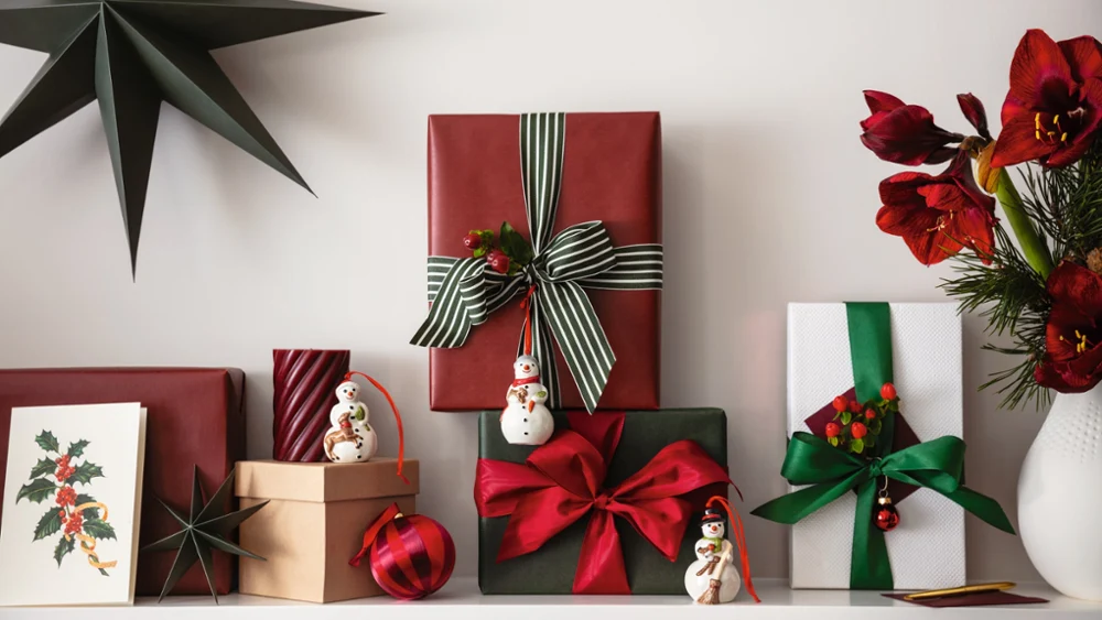 Christmas presents on a mantelpiece A charming arrangement with nostalgic ornaments from Villeroy & Boch, including wrapped gifts with bows, a bright red star ornament, adorable snowman figurines and a vase of lush red flowers.