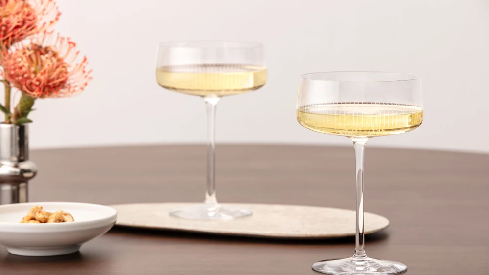 Twee Afina glazen gevuld met mousserende wijn op een houten tafel, met bloemen en een kleine schaal ernaast.