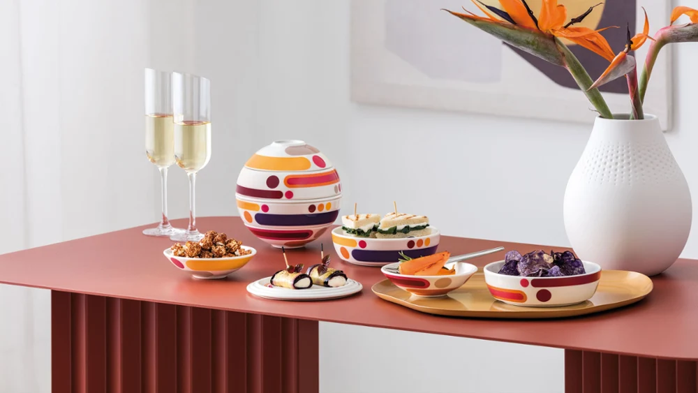 Moderne tafelsetting met La Boule Miami, diverse hapjes, twee champagneglazen en een witte Manufacture Collier vaas met oranje bloemen.