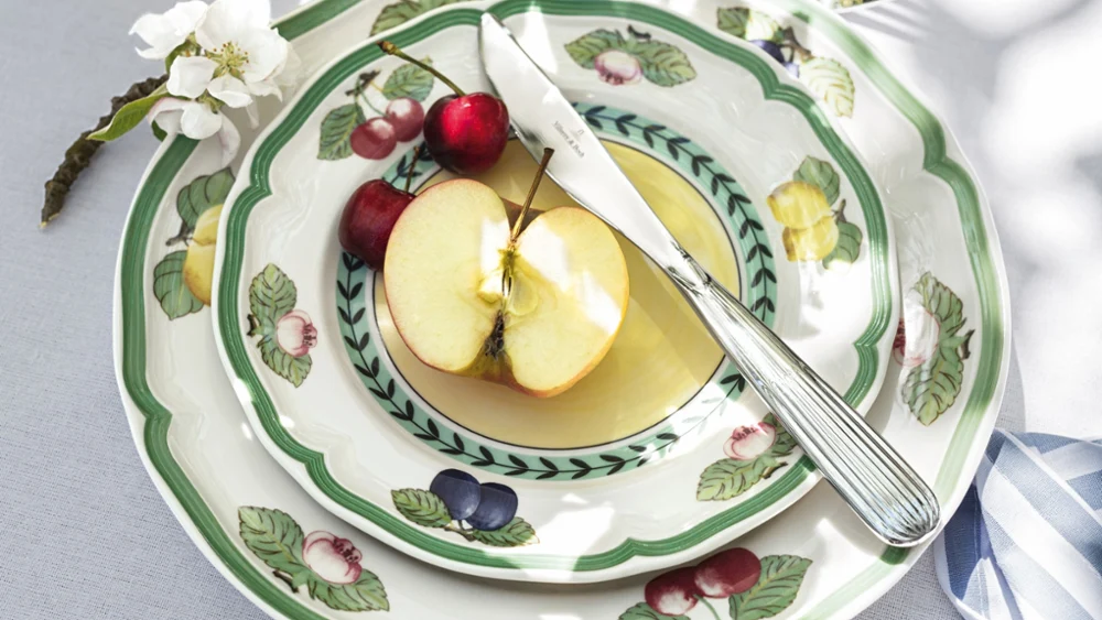 A table setting with stacked Villeroy & Boch French Garden floral-patterned plates, along with a sliced apple, two cherries, a knife and a fork. Next to them are white flowers and a blue ribbon.